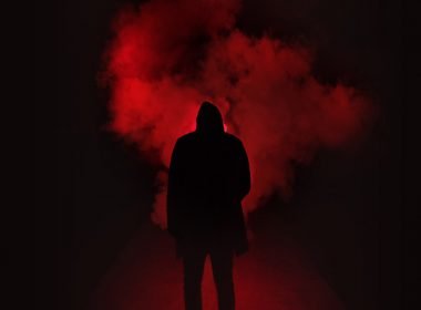 A dark hooded figure at night stands in a foggy street illuminated with a dim red light.