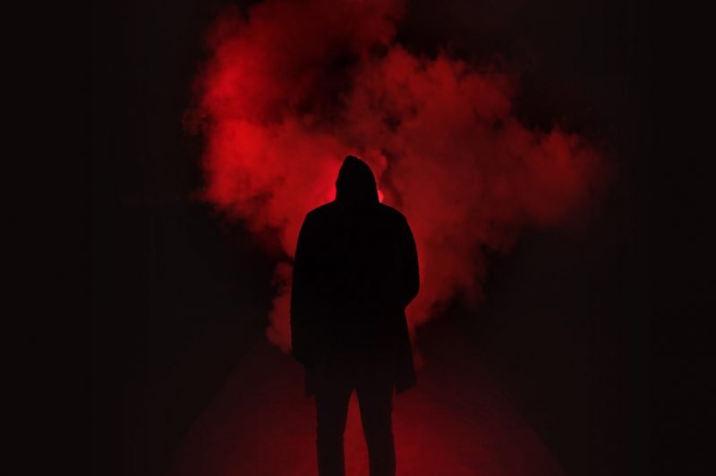 A dark hooded figure at night stands in a foggy street illuminated with a dim red light.