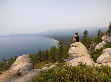 A hiker sits atop the head of Monkey Rock overlooking the large Lake Tahoe and groups of swimmers enjoying the water below.