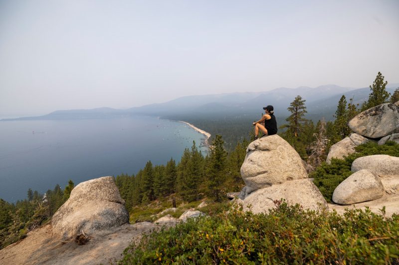 A hiker sits atop the head of Monkey Rock overlooking the large Lake Tahoe and groups of swimmers enjoying the water below.