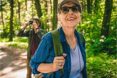 A woman hiking through the forest in the spring while her partner uses binoculars to bird watch behind her.