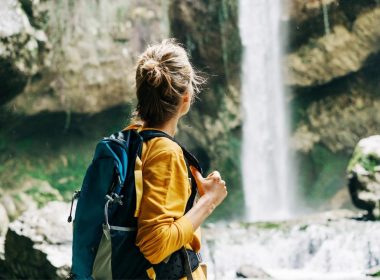 From behind, a woman with her hair in a bun and a hiking pack views a tall waterfall crashing over rocks in the forest.