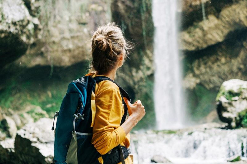 From behind, a woman with her hair in a bun and a hiking pack views a tall waterfall crashing over rocks in the forest.