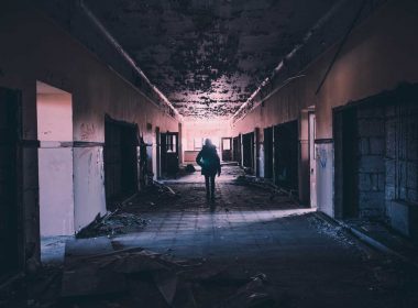 A woman explores down a dark hallway of an abandoned building with moss growing on the ceiling.