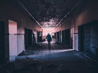 A woman explores down a dark hallway of an abandoned building with moss growing on the ceiling.