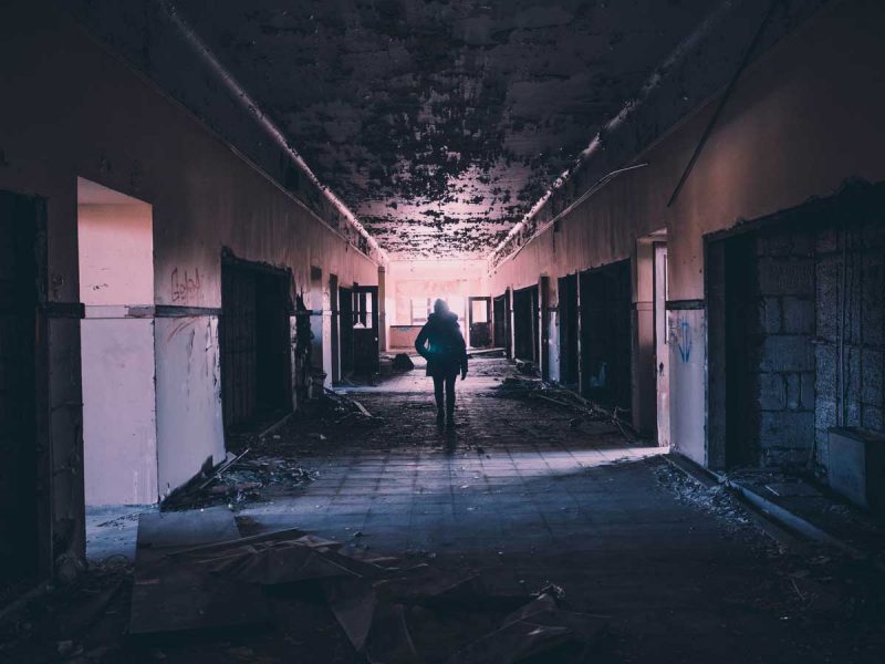 A woman explores down a dark hallway of an abandoned building with moss growing on the ceiling.