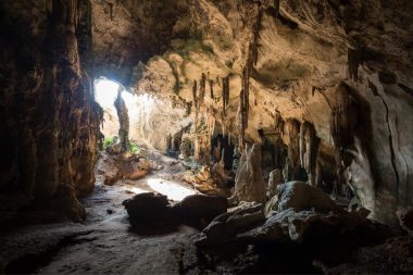 An underground cave with light filtering in from the mouth has stalactite and stalagmite on the ceiling and ground.