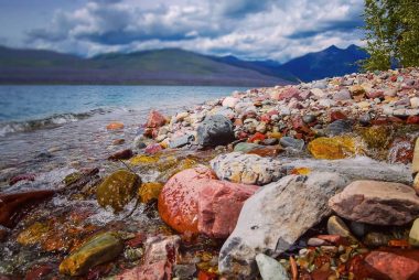 The colorful rocky shoreline of Lake McDonald with tall blue mountains across the lake.