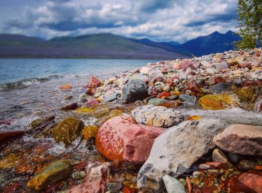 The colorful rocky shoreline of Lake McDonald with tall blue mountains across the lake.