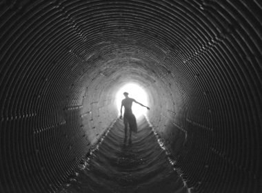A silhoutte of a man walking down a ridged culvert tube below a road.