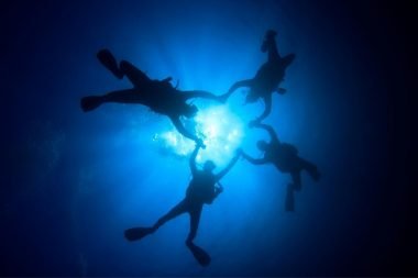 From below, four scuba divers hold hands to create a clover shape in the water as they dive.