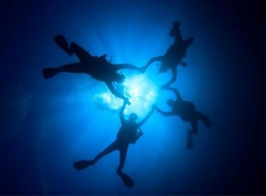From below, four scuba divers hold hands to create a clover shape in the water as they dive.