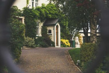Looking through a black, link fence to an old wooden house with a gravel driveway.