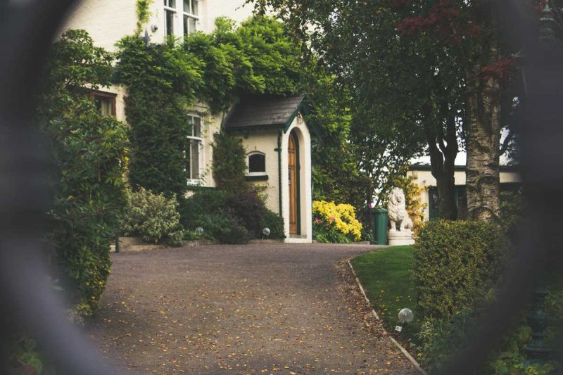 Looking through a black, link fence to an old wooden house with a gravel driveway.