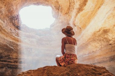 A woman sits on a boulder inside a massive cave with a tall ceiling, looking up at the light pouring in through the entrance above.