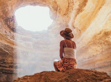 A woman sits on a boulder inside a massive cave with a tall ceiling, looking up at the light pouring in through the entrance above.