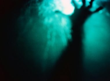 The dark form of a blurry hand under dark water reaching towards the light at the surface of the water.