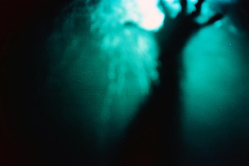 The dark form of a blurry hand under dark water reaching towards the light at the surface of the water.