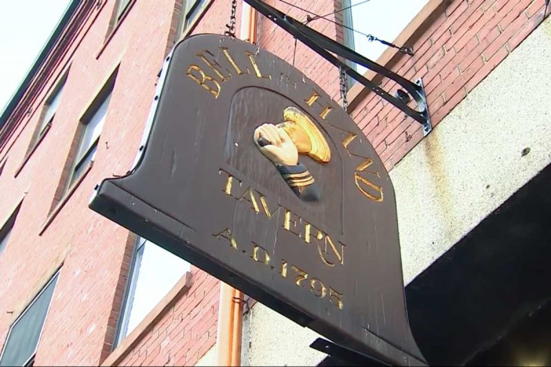 A brown wooden sign hanging outside Bell In Hand Tavern in Boston.