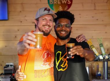 Two bros holding up craft beers and smiling in a brewery.