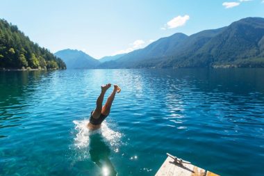 A man diving into the lake with his feet sticking out of the beautiful blue water surrounded by tree covered mountains.