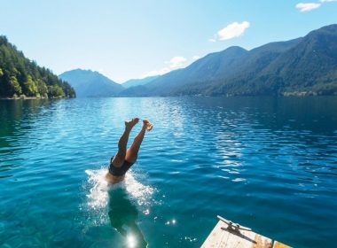 A man diving into the lake with his feet sticking out of the beautiful blue water surrounded by tree covered mountains.