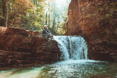 A hiker sits on a cliff edge near a waterfall as sun filters through the canopy above.