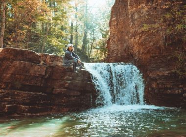 A hiker sits on a cliff edge near a waterfall as sun filters through the canopy above.