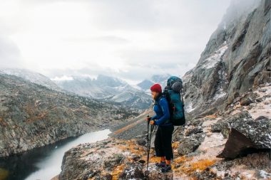 A backpacker stands with hiking poles on rocky terrain overlooking the Wind River Range Mountains and a river in Wyoming