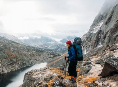 A backpacker stands with hiking poles on rocky terrain overlooking the Wind River Range Mountains and a river in Wyoming