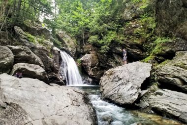 A hiker stands on a boulder in the forest and looks out across a creek to Bingham Falls in Vermont.