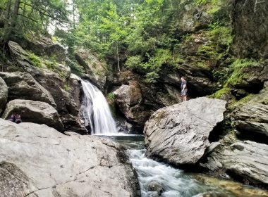 A hiker stands on a boulder in the forest and looks out across a creek to Bingham Falls in Vermont.