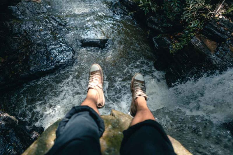 A woman hangs her feet over the edge of a waterfall as she sits on a boulder.
