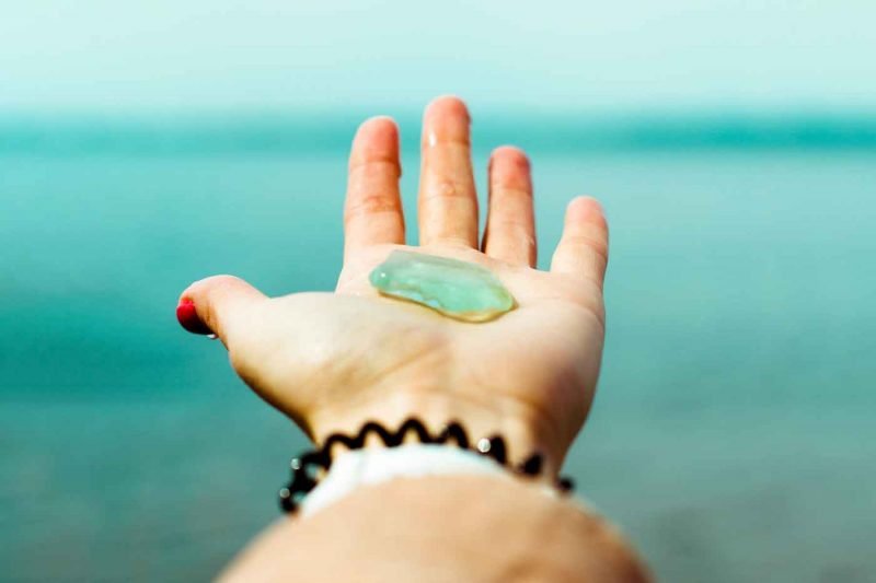 Holding up a piece of smooth aqua sea glass matching the color of the ocean water in the distance.