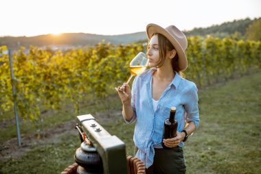 Young and cheerful woman tasting wine, while standing near the press machine on the vineyard on a sunny evening