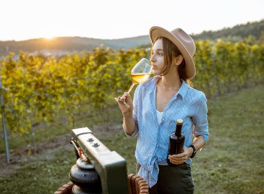Young and cheerful woman tasting wine, while standing near the press machine on the vineyard on a sunny evening