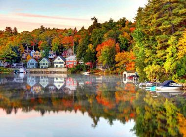 Colorful fall leaves and colorful homes and business buildings along the shoreline of Lake Winnipesaukee, New Hampshire, reflecting on the water in the morning light.