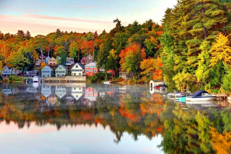 Colorful fall leaves and colorful homes and business buildings along the shoreline of Lake Winnipesaukee, New Hampshire, reflecting on the water in the morning light.